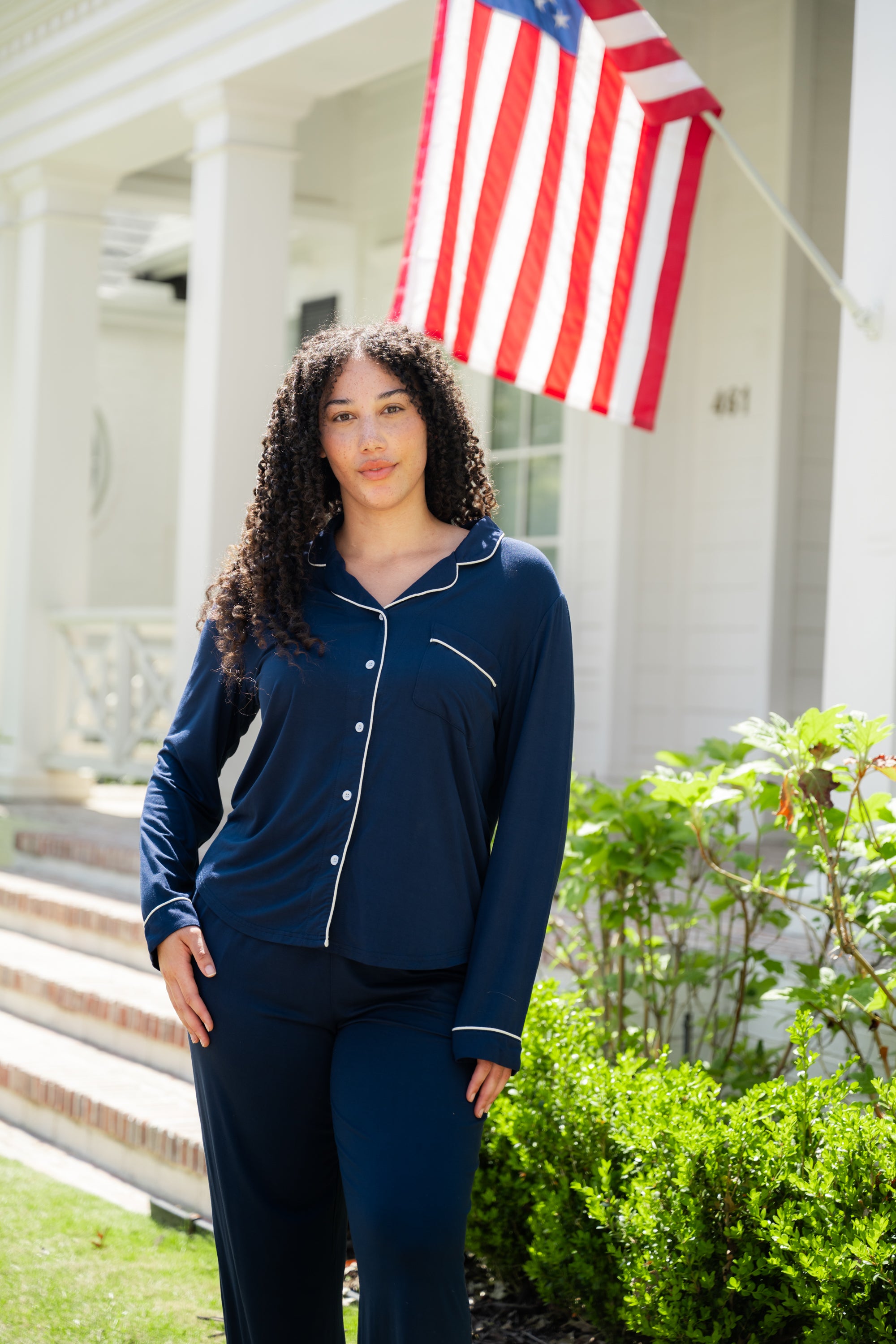 Woman wearing a navy blue pajama set standing in front of a house with an American flag.