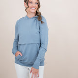 Smiling female model wearing the Women's French Terry Nursing Hoodie in Slate with one hand in the kangaroo pocket paired with white pants