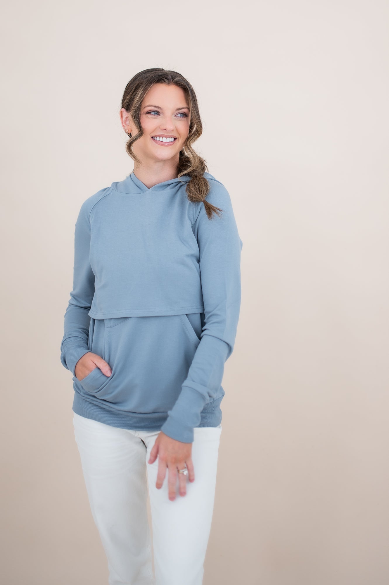 Smiling female model wearing the Women's French Terry Nursing Hoodie in Slate with one hand in the kangaroo pocket paired with white pants