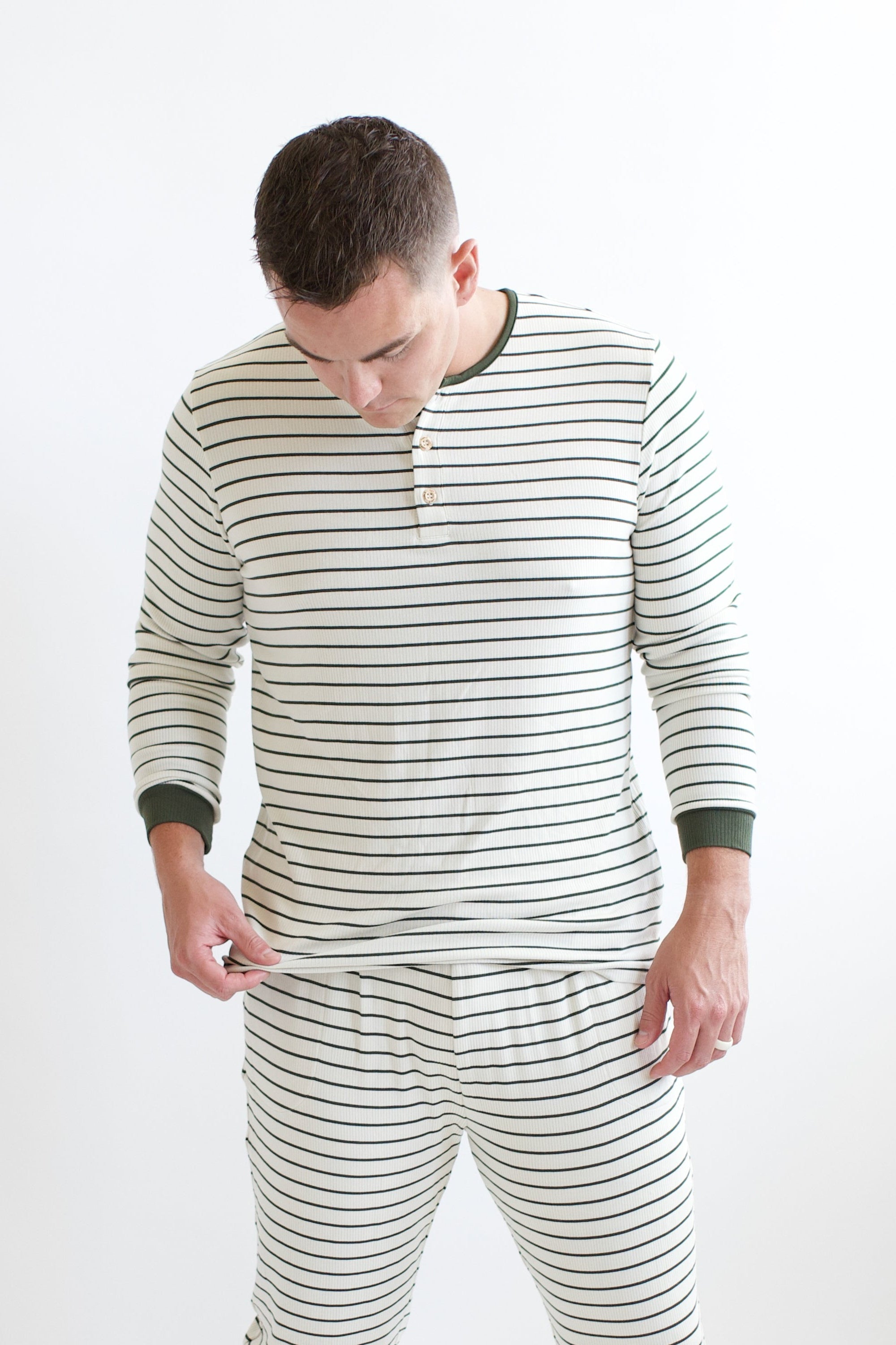 Male model holding the hem of the shirt of the Men's Ribbed Henley Set in Fir Stripe in front of a white background
