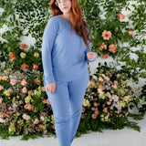 Woman modeling Women's Jogger Pajama Set in Periwinkle color with a wall of greenery behind 