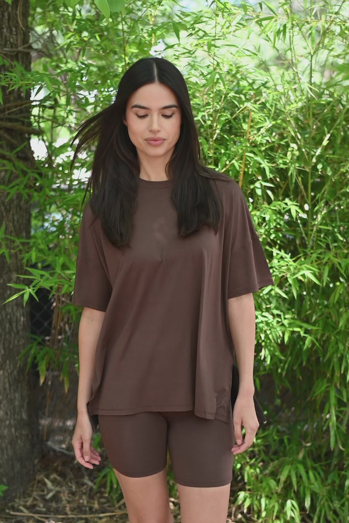 Video of Woman in brown oversized t-shirt and biker shorts standing outdoors with a white fence and trees in the background. ::: Model is 5'10" wearing size S ///