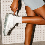 Person wearing light blue shorts and white sneakers with socks with a patterned wall in the background